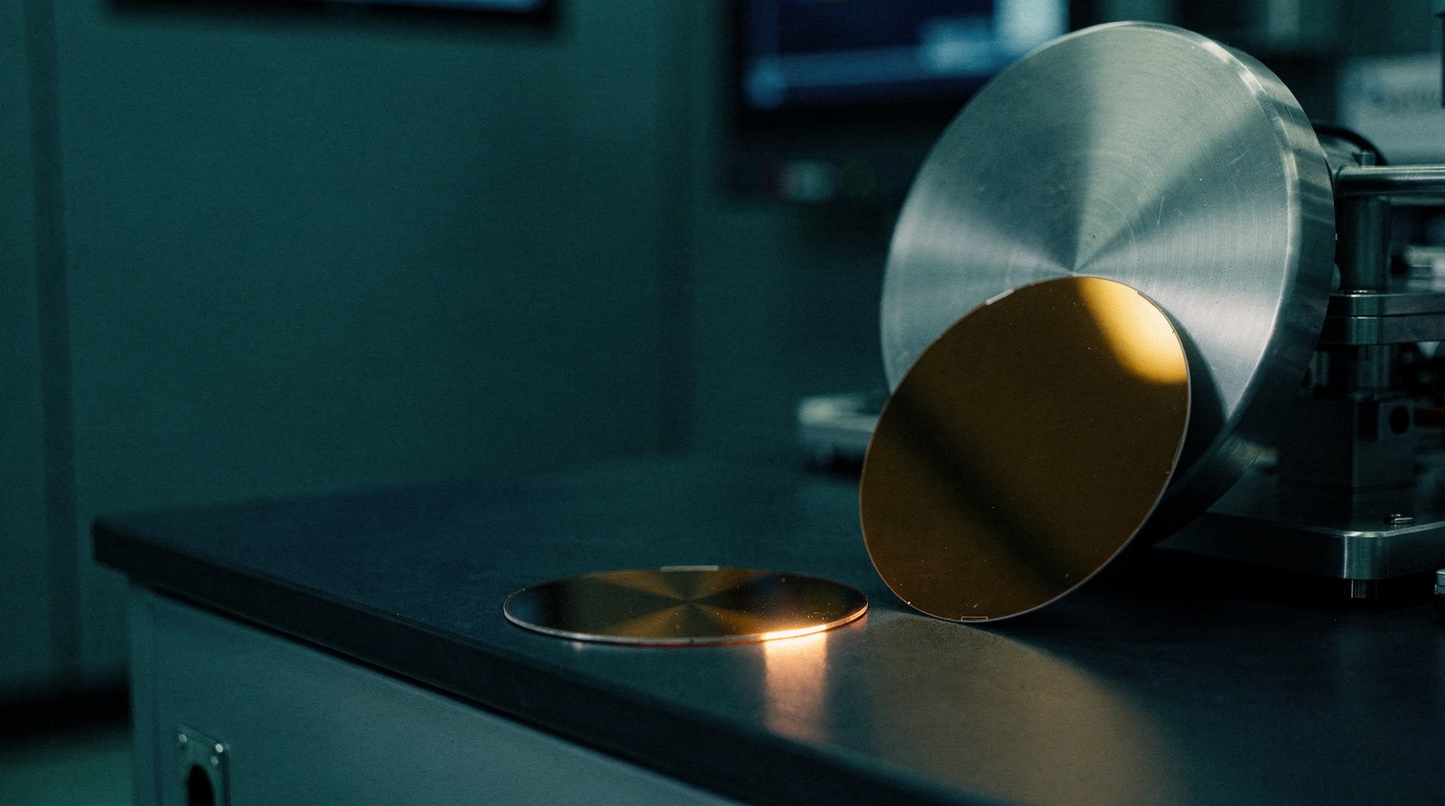 A small six-inch wafer beside a large 300mm wafer on a dark bench, the small wafer lit warm gold